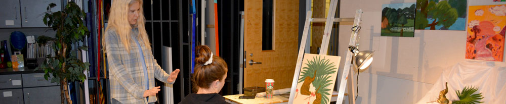Student sitting at a painting on an easel and a teacher speaking with the student