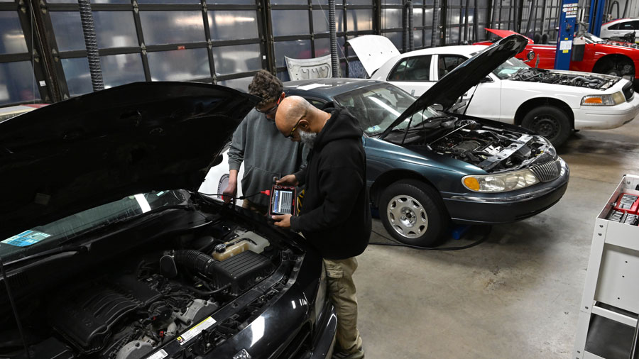 Two students using a diagnostic tool on a car with the hood up