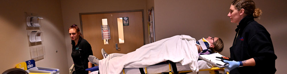 A student on a stretcher with two other EMT students manuvering it throught a hallway