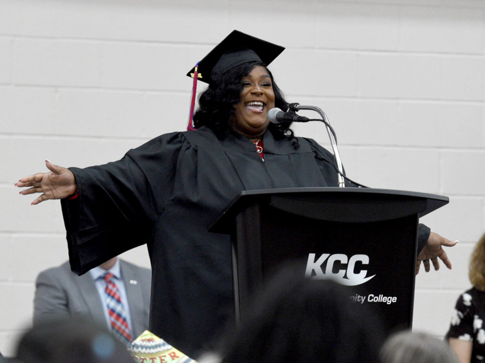 A smiling graduate in a cap and gown at a KCC podium with arms wide during a graduation ceremony.