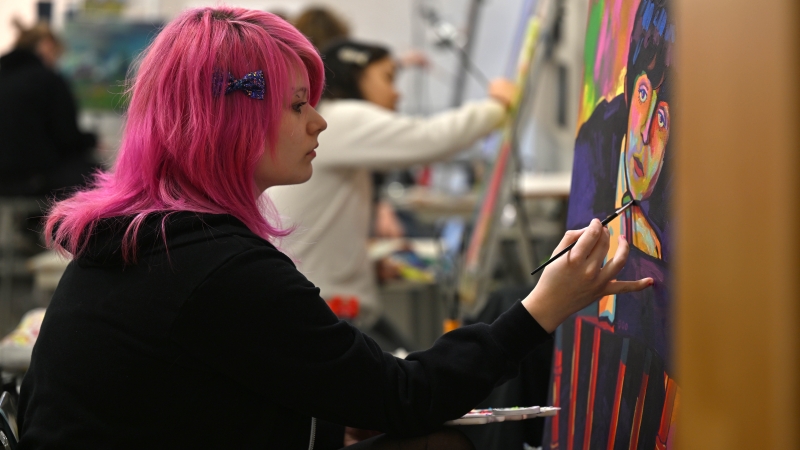 Student with bright pink hair painting a colorful portrait in an art studio.