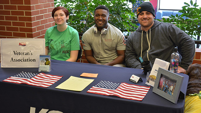 Photo of three members of the Veterans Association (and a service dog) siting at their booth at 'Club Rush'