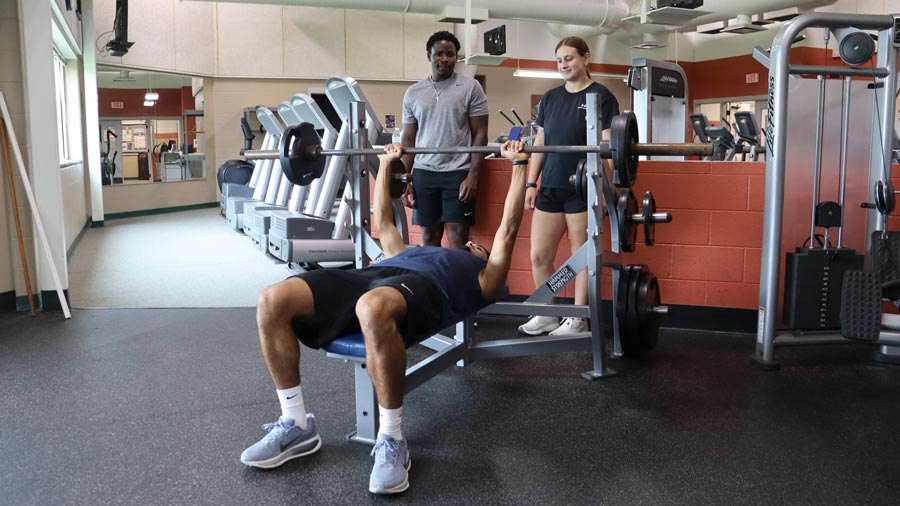 Student bench pressing weights at the KCC Fitness Center with two spotters.